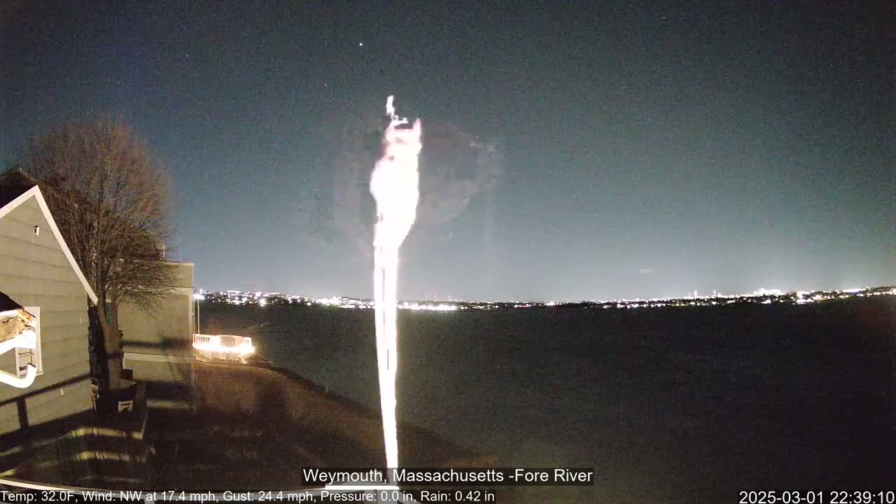 A bright light, possibly a searchlight or similar, shines upward into the night sky over a dark body of water, with a house and lights visible on the shore under a partly cloudy sky.