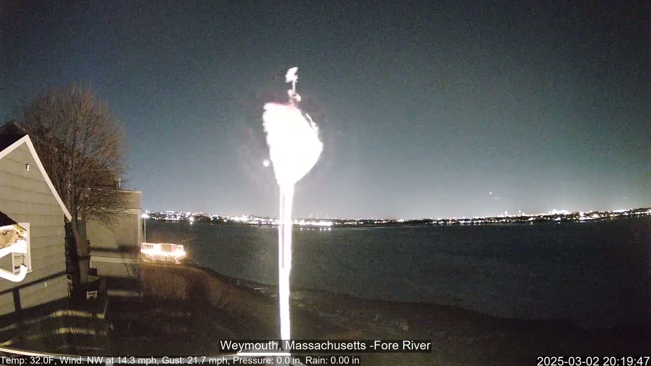 A bright light, possibly a flare, rises from a body of water at night under a clear sky, with a house and a dark tree visible in the foreground.