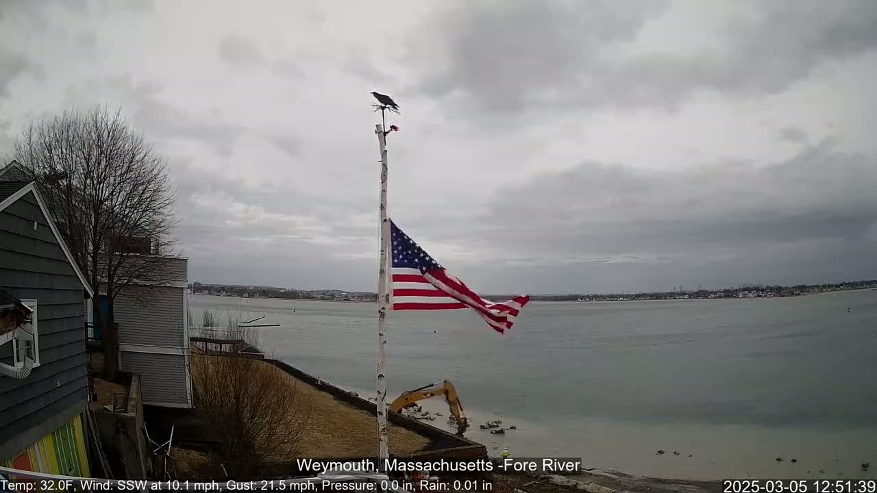 An American flag flies from a birch pole near a grey house overlooking a grey, calm river under an overcast sky.