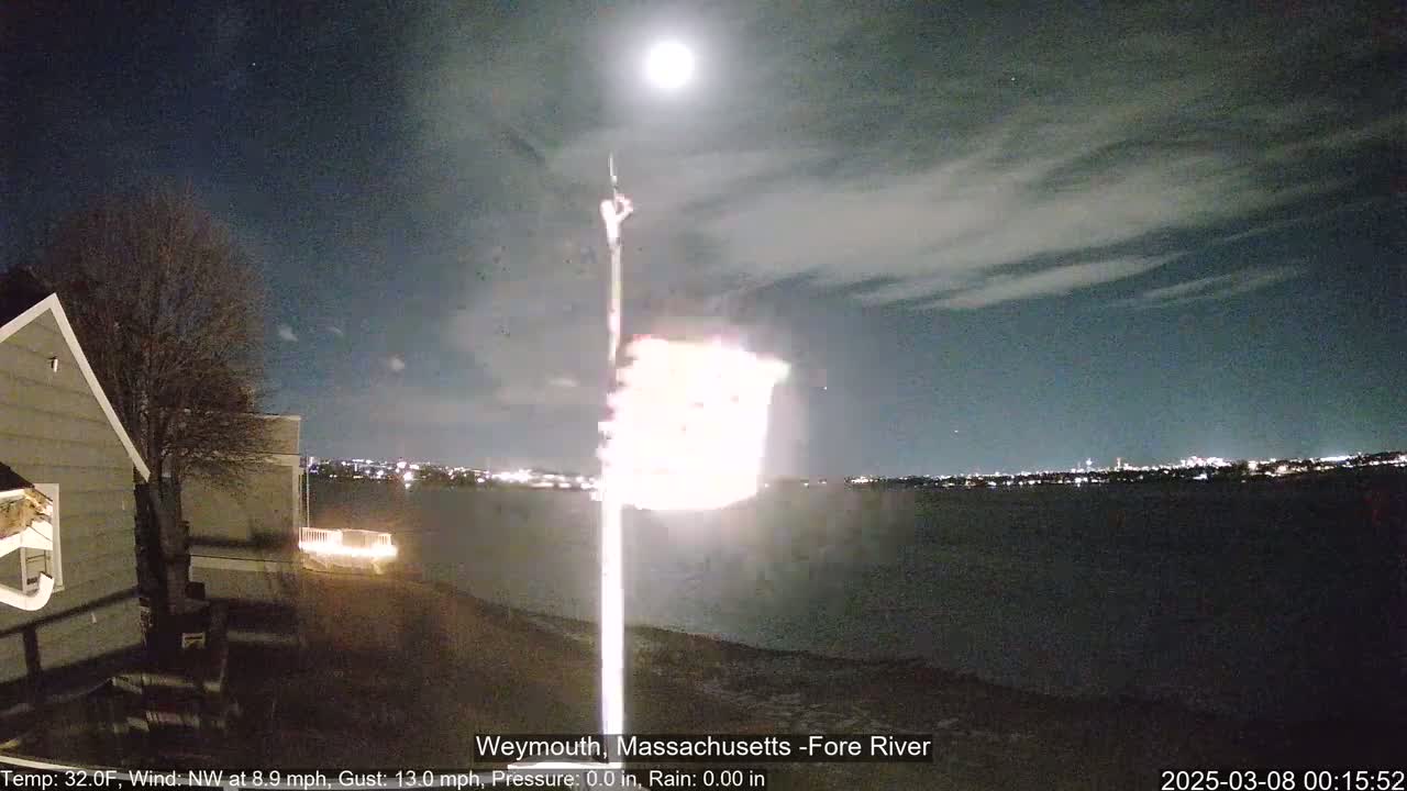A nighttime view of a body of water under a partly cloudy sky and bright moon, with a flag illuminated on a pole in the foreground and a house partially visible to the left.