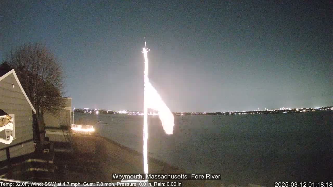 A nighttime view of a body of water with a flagpole and a house visible in the foreground under a dark, clear sky.