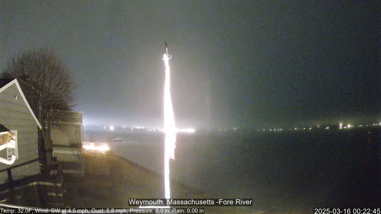 A brightly lit flagpole stands against a foggy, dark nighttime sky over a body of water with distant lights.