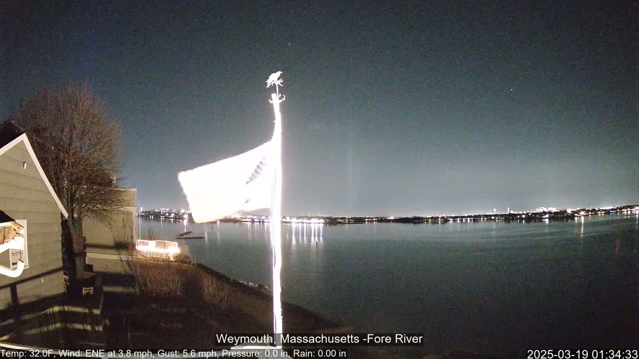 A nighttime view of a calm body of water reflecting city lights across the horizon, with a flag waving on a pole near a house under a clear, dark sky.