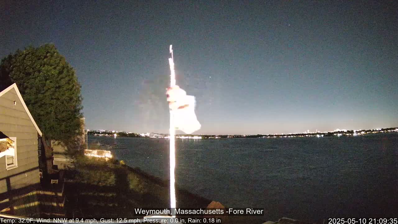 A nighttime view of a body of water with city lights across the way, a flagpole with a blurred flag, and a house to the left under a clear, dark sky.