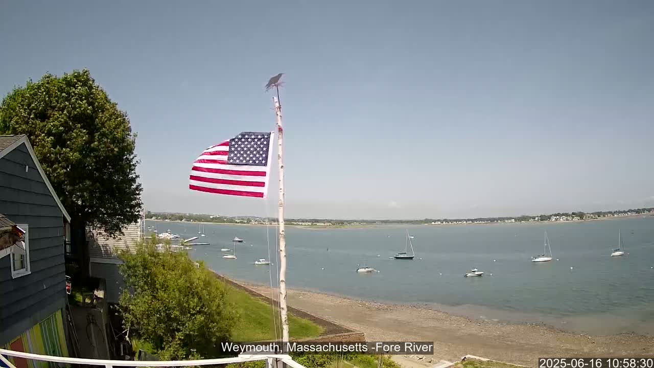 An American flag flies on a pole beside a house overlooking a calm bay with several sailboats and motorboats, under a clear sky.