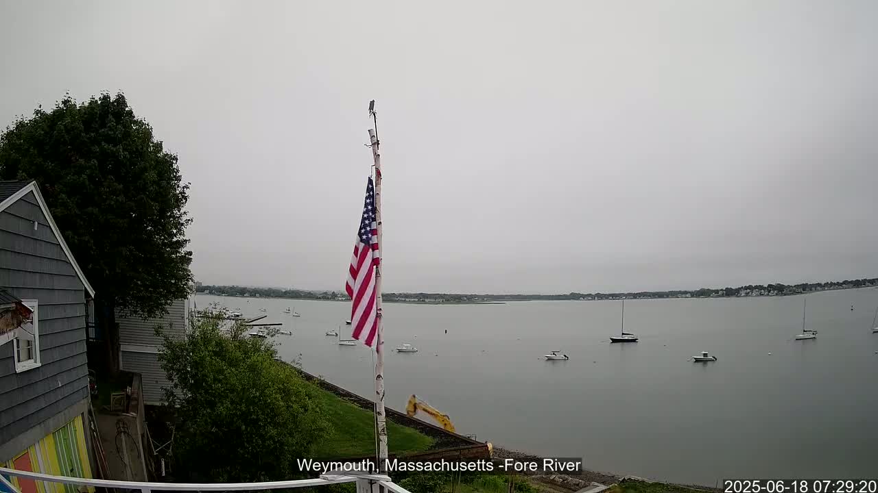 A gray, overcast day reveals a calm body of water dotted with several sailboats and motorboats, viewed from a hillside with a house and an American flag on a pole.