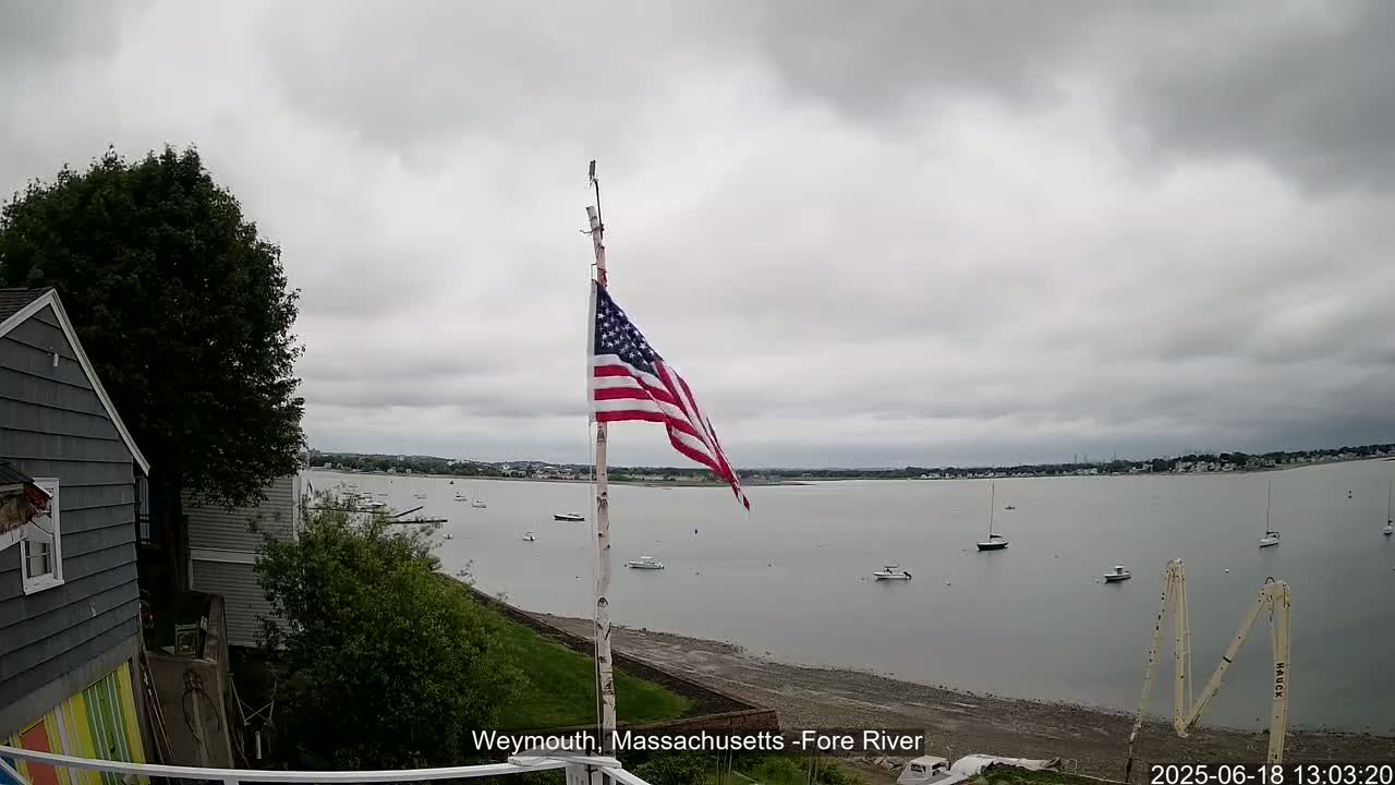 An American flag flies on a pole near a gray house overlooking a calm, overcast body of water with several small boats.