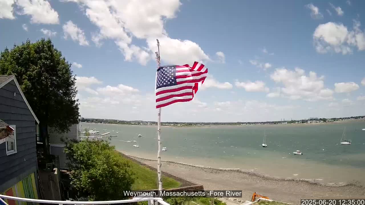 An American flag waves on a pole overlooking a calm, partly cloudy day at a waterfront with several boats and a sandy shore.