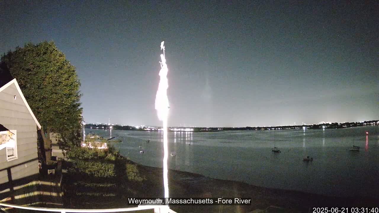 A nighttime view from a backyard shows a calm body of water with a few boats, a house to the left, and a flagpole in the center under a clear, dark sky.