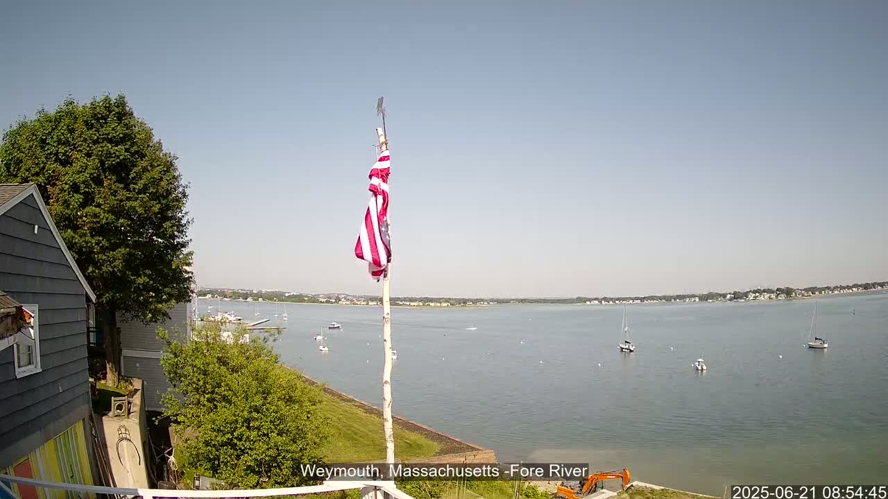 A partly cloudy sky overlooks a calm body of water with several sailboats, a house and a  flagpole with an American flag on it, all situated on a grassy hill.
