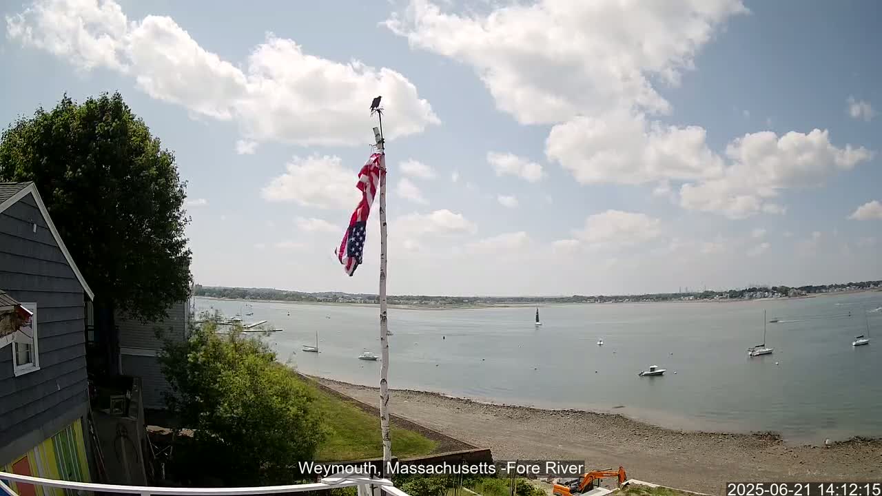 A partly cloudy day shows a view of a calm body of water with several boats, a sandy shore, and a house with a flagpole displaying an American flag.
