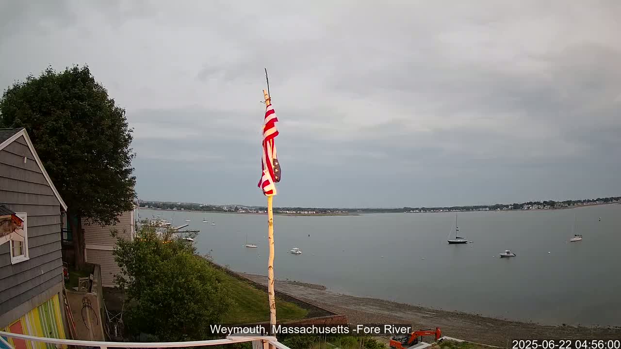 A gray house with a colorful lower section sits on a hill overlooking a calm, gray body of water with several boats, under an overcast sky, and a slightly tattered American flag is flown on a pole in the foreground.