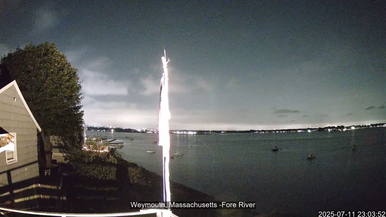 A nighttime view shows a dark body of water with several boats, a house partially visible on the left, and a bright light source extending across the image from left to right under a dark, overcast sky.