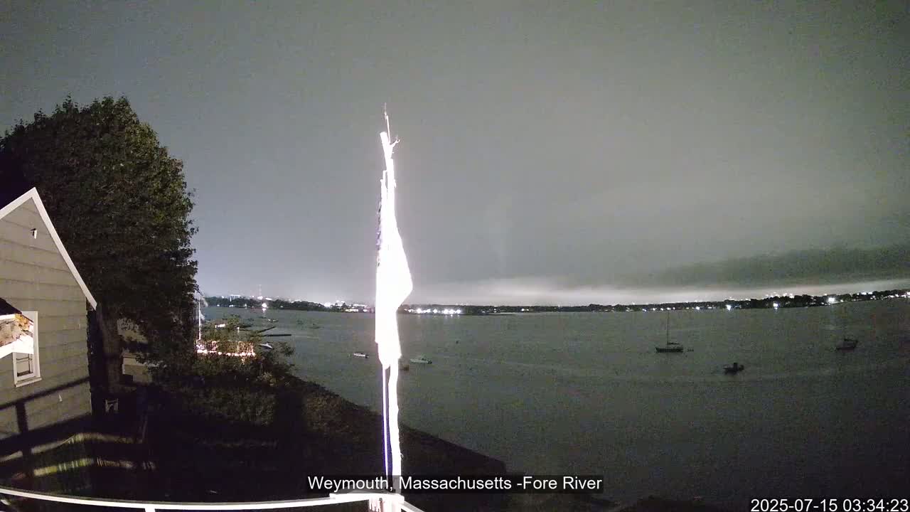 A nighttime view of a calm body of water with a few boats, seen from a backyard with a house and a flagpole partially visible under an overcast sky.