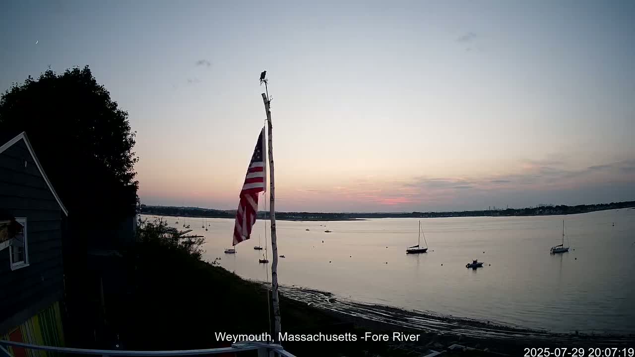 An American flag flies on a pole overlooking a calm body of water with several boats, under a mostly sunny sky.