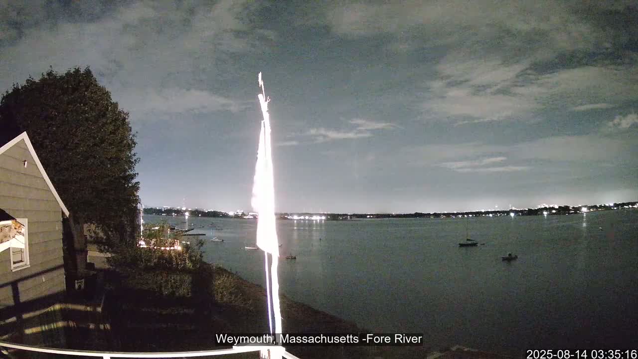 A nighttime view shows a calm body of water with a few boats, a house partially visible on the left, and a flagpole in the foreground under a cloudy sky.