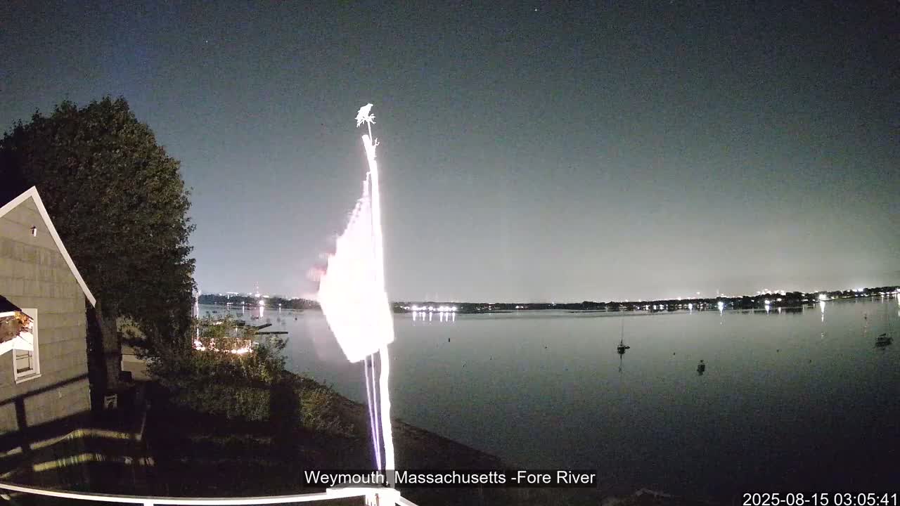 A nighttime view shows a calm body of water with a few boats, a house and trees to the left, and a flag illuminated by an unseen light source.