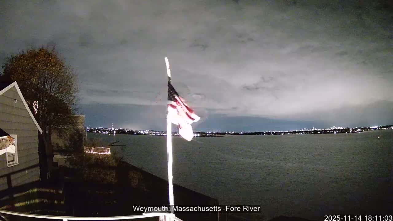 An illuminated American flag stands on a pole next to a house overlooking a calm body of water with a distant city skyline under a dark, heavily overcast night sky.