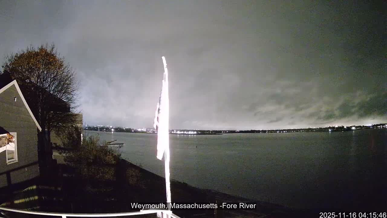 On an overcast night, a brightly lit flag stands prominently on a pole at the water's edge, with a house and trees to the left, and distant city lights reflected across the dark water.