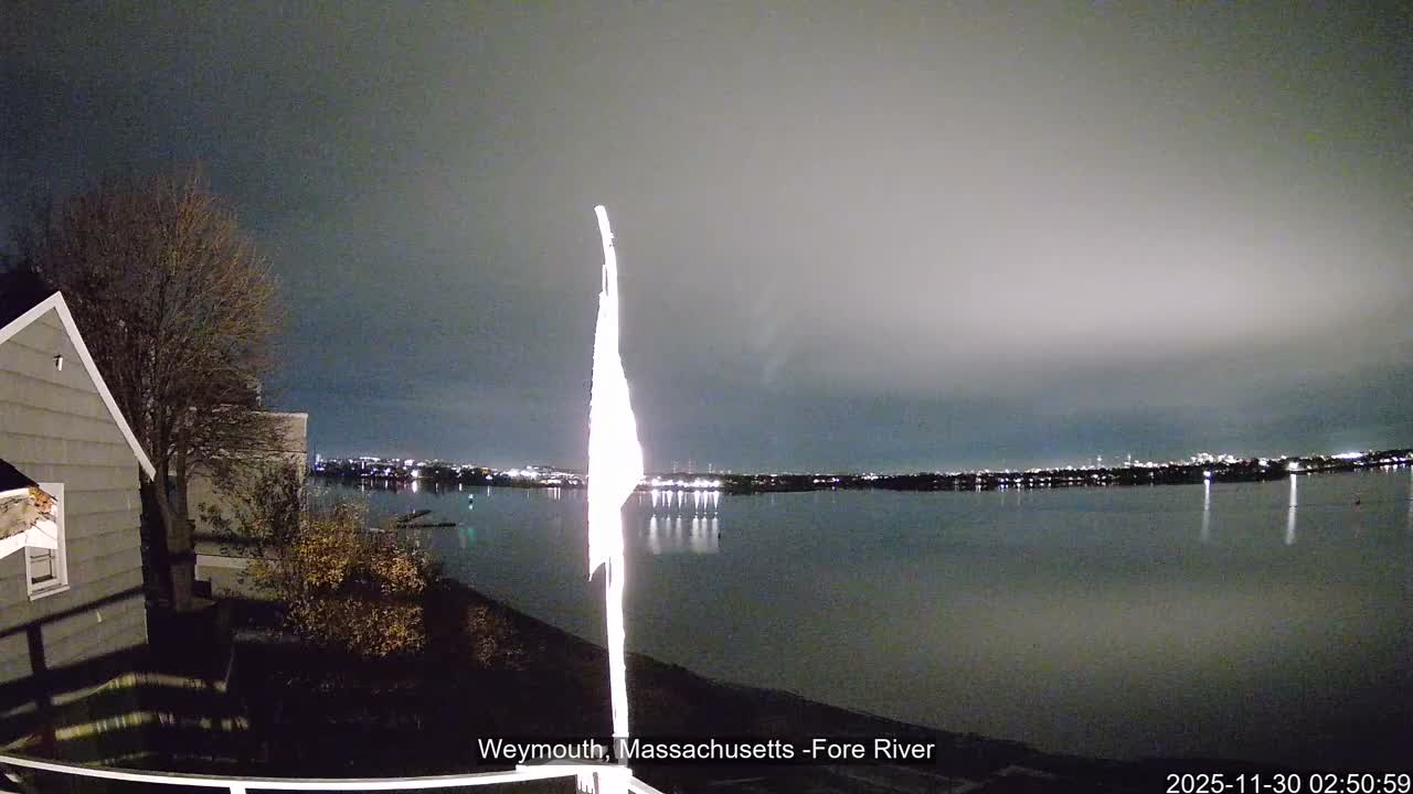 A dark, overcast night scene captures a wide river or bay with a brightly illuminated vertical structure reflecting on the water, flanked by a house and bare trees on the left, and a distant illuminated cityscape across the water.