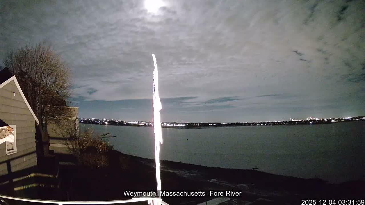 A cloudy night scene reveals a bright moon illuminating a river and a distant city skyline, with a brilliantly lit flag, a house, and a bare tree occupying the foreground.