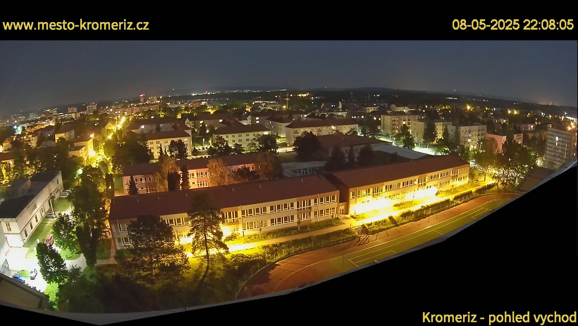 A nighttime aerial view shows a town with illuminated buildings and streets, including a large school with a running track, under a clear, dark sky.