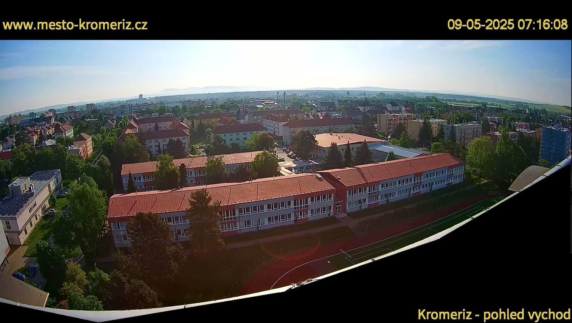 An aerial view on a sunny day shows a large school complex with red roofs and surrounding buildings in a town, next to a running track and playing field.