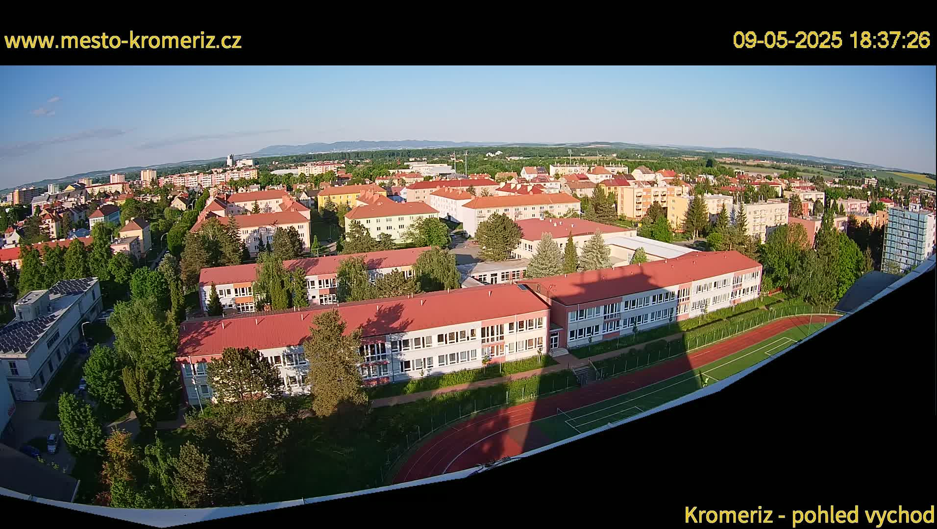 A high-angle, long shot shows a sprawling town under a clear, sunny sky, featuring numerous low-rise buildings surrounded by lush green trees and a running track.