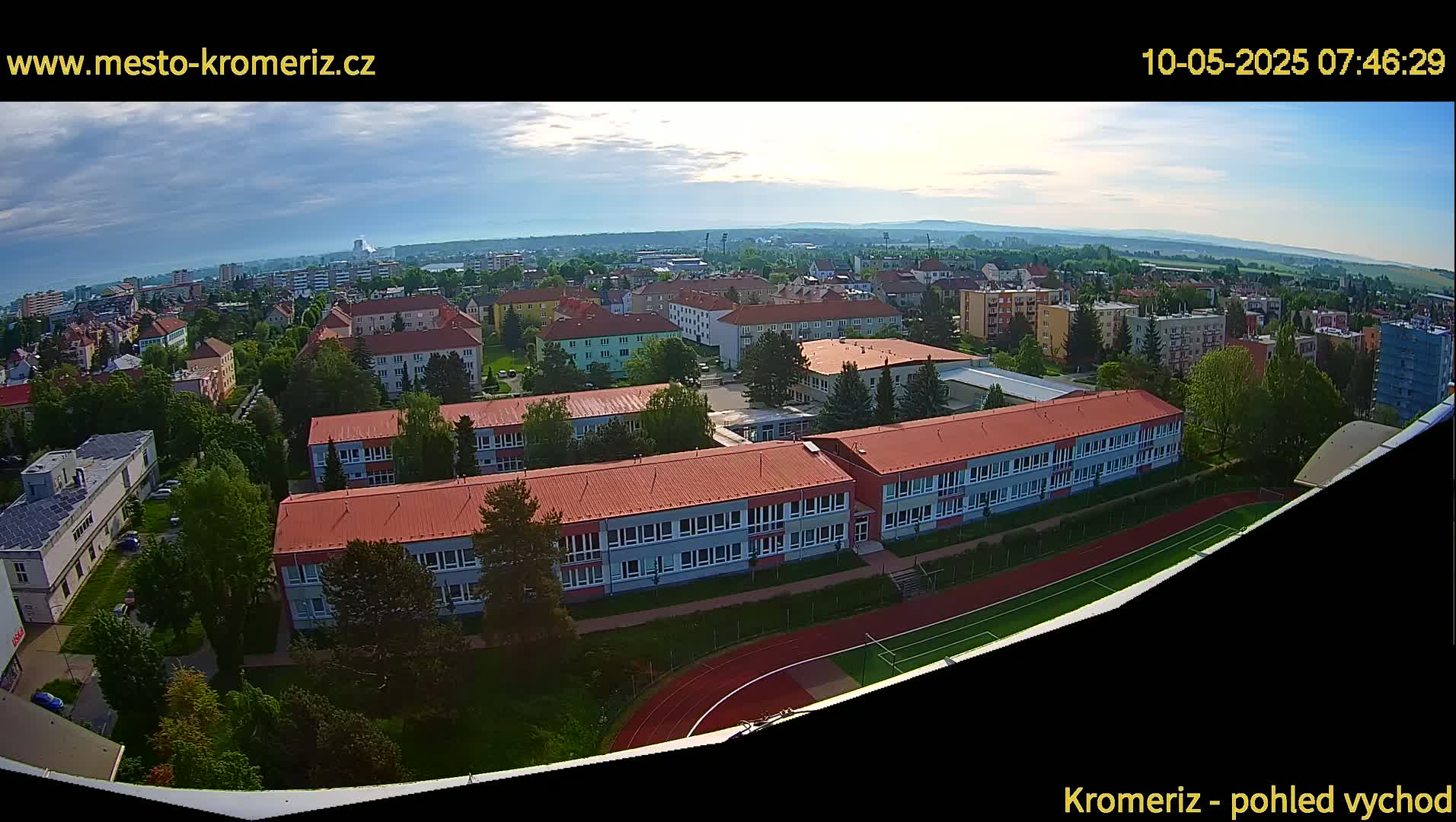 An aerial view shows a large school building with red roofs and a running track, surrounded by other buildings and green trees under a partly cloudy sky.