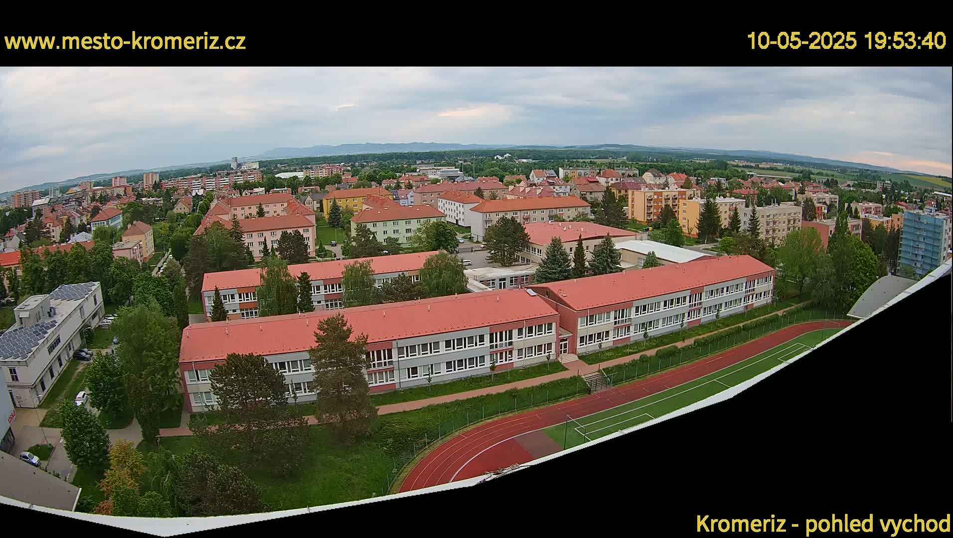 An aerial view shows a school and surrounding residential buildings on a partly cloudy day, with a running track and playing field visible next to the school.