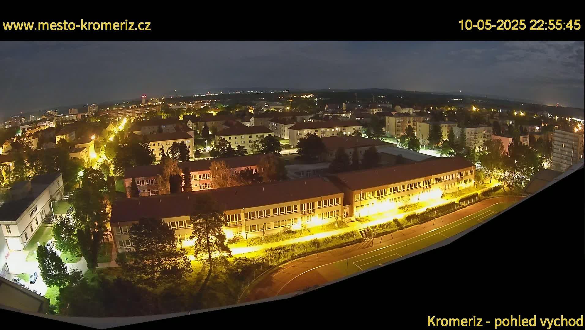 A nighttime aerial view shows a town's buildings and streets illuminated, with a running track visible in the foreground under a clear, dark sky.