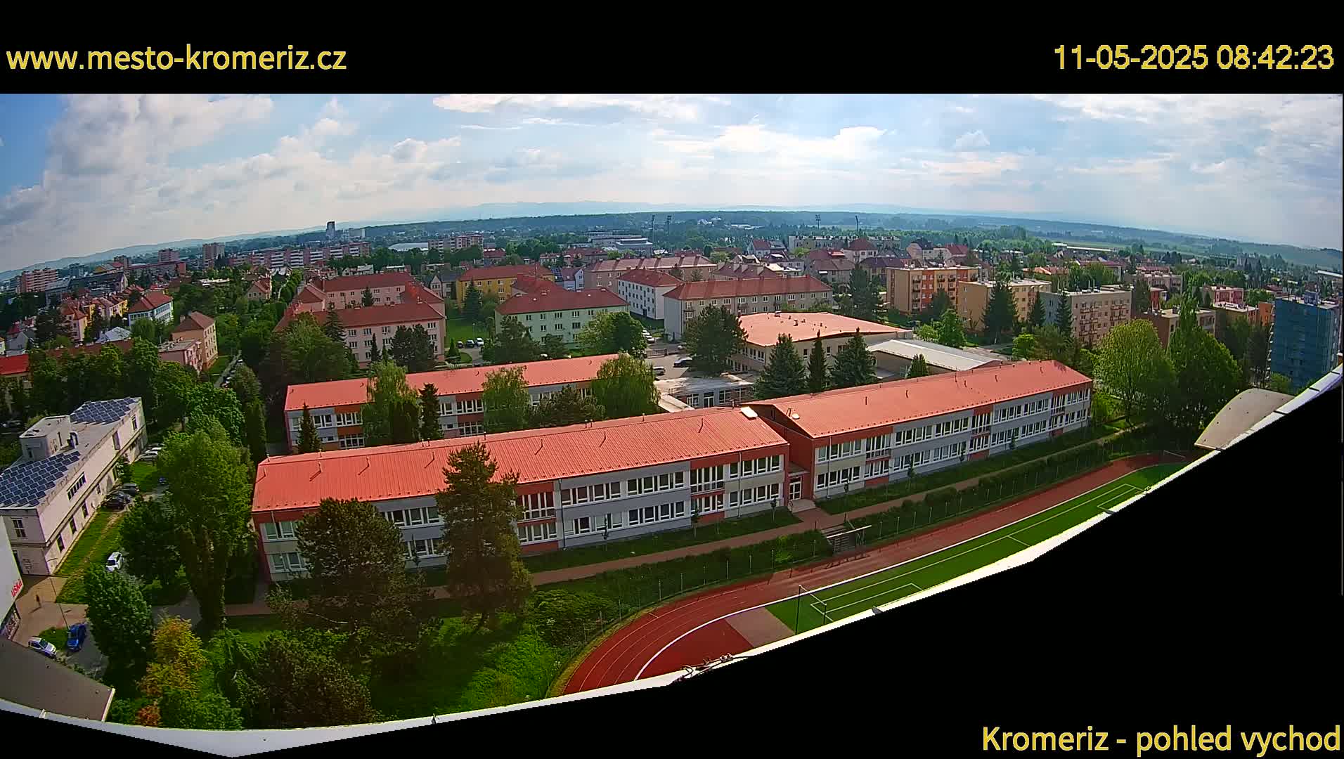 A partly cloudy day reveals a high-angle view of a school campus with red-roofed buildings, a running track, and a soccer field, nestled within a residential area and surrounded by lush green trees.
