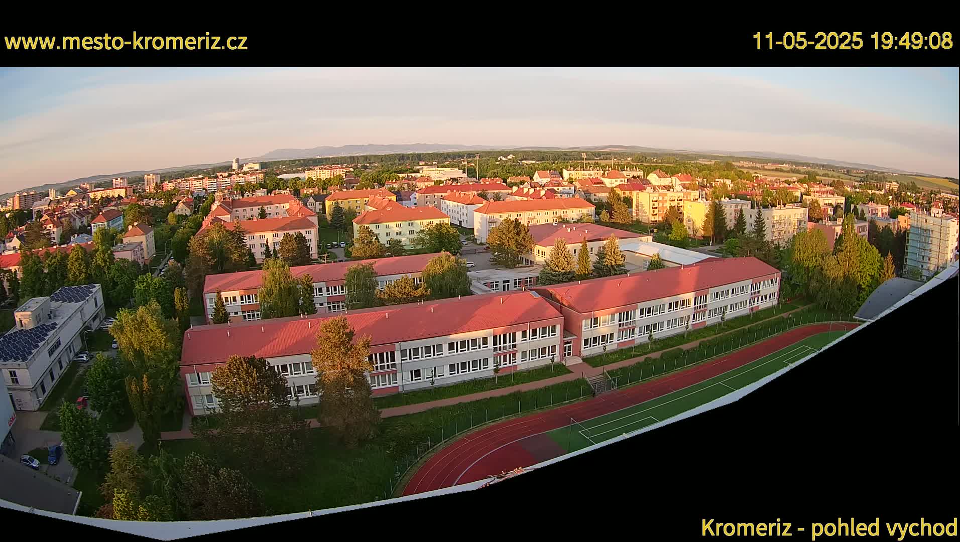 An aerial view shows a residential area with a large school and running track, bathed in the warm light of a setting sun.