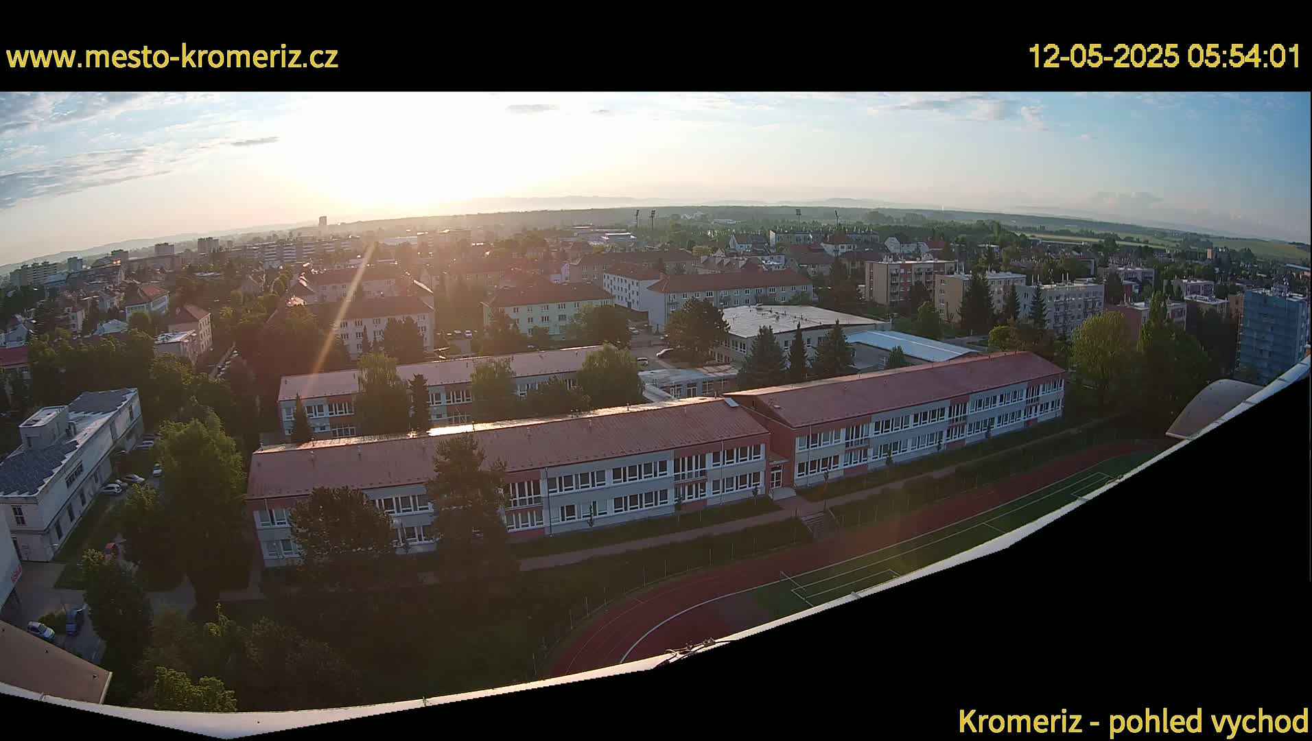 A high-angle, sunrise view of a town with a large school building and athletic field, surrounded by residential buildings and trees.