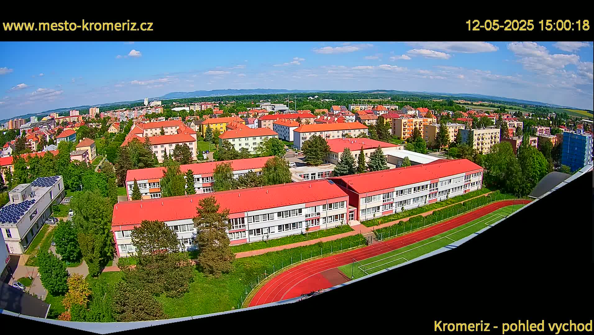 An aerial view shows a town on a sunny day with many low-rise buildings, green spaces, and a red running track.