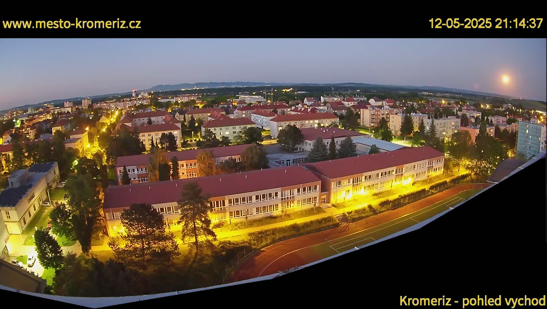 An aerial view at twilight shows a town with illuminated buildings and a running track, under a mostly clear sky.
