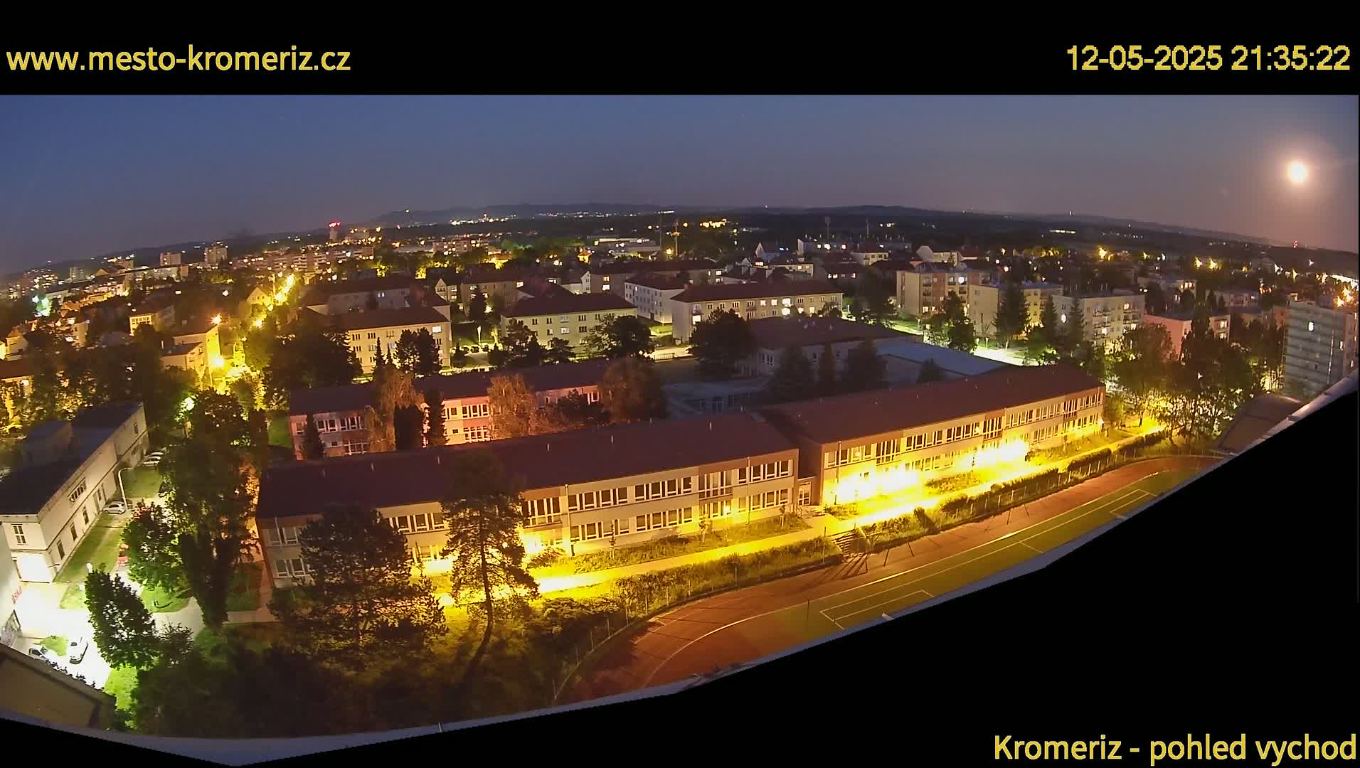 A nighttime aerial view of a town shows illuminated buildings and streets, with a running track visible in the foreground under a clear, moonlit sky.