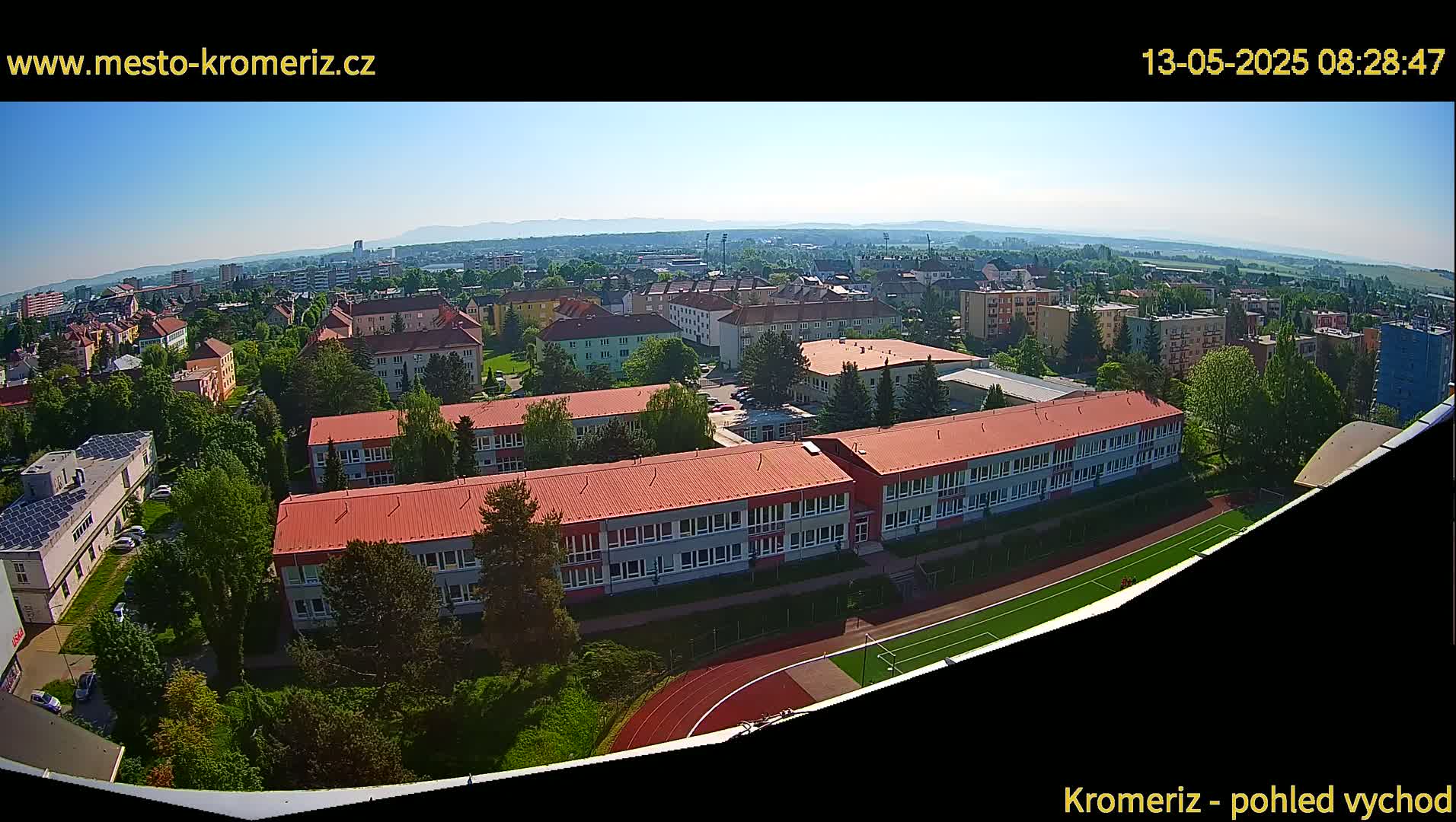 A high-angle, sunny view shows a large school complex with red roofs and surrounding buildings and greenery, next to a running track and field.