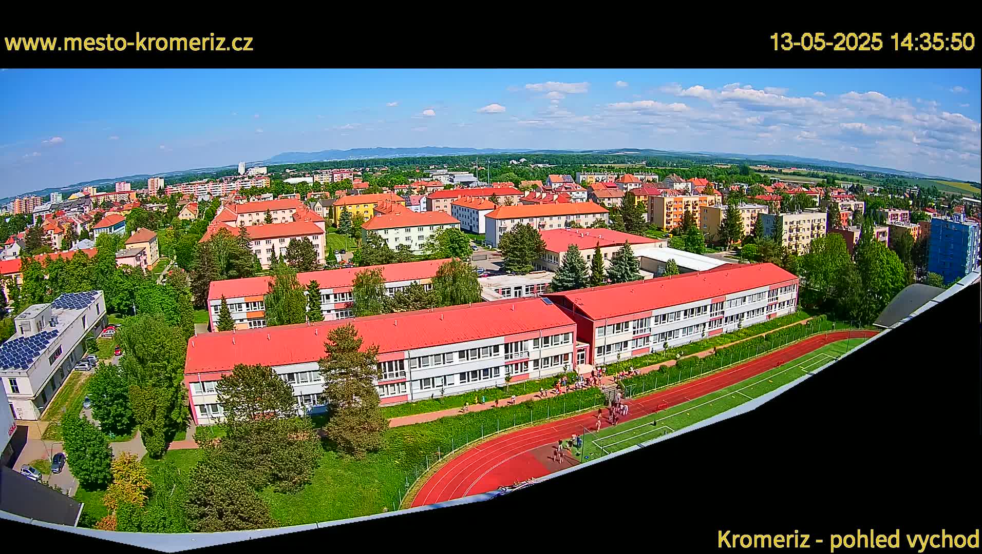 An aerial view shows a large school with red roofs and a running track, situated amongst a residential area on a sunny day.