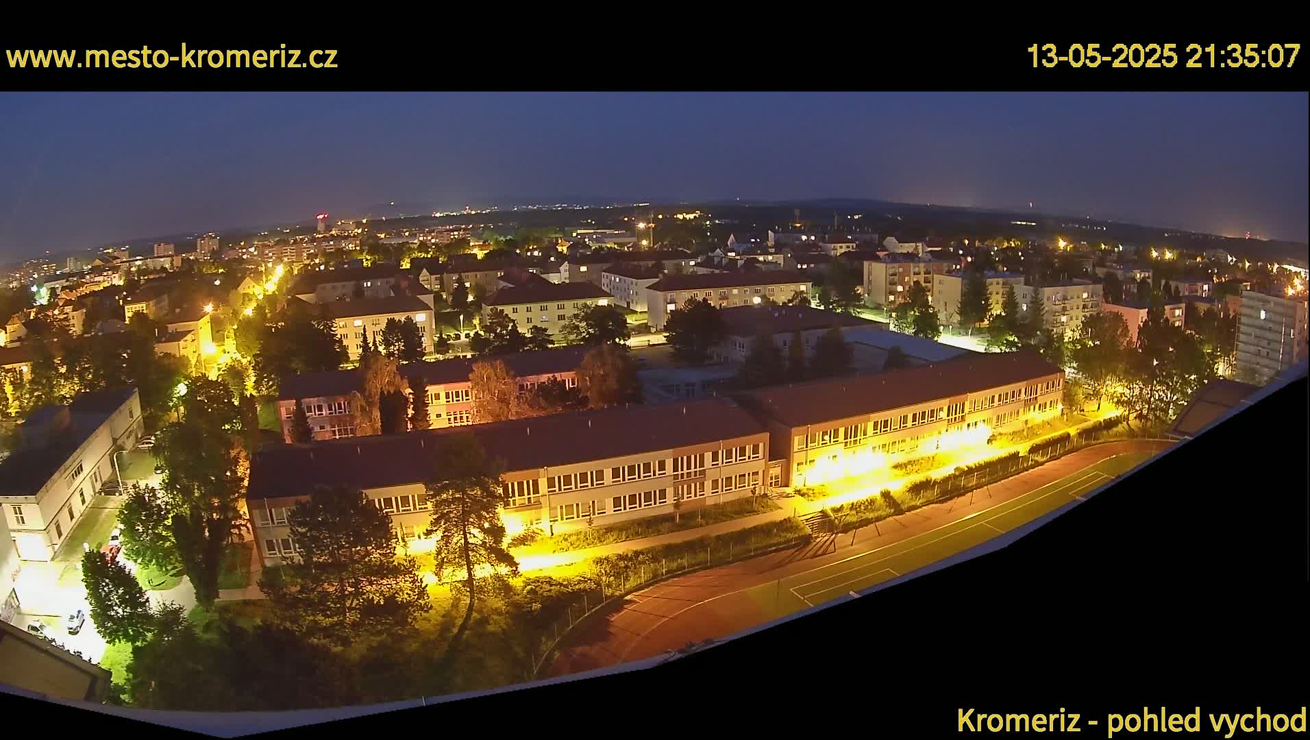 A nighttime aerial view of a town shows buildings illuminated with streetlights and a school with a running track and playing field.