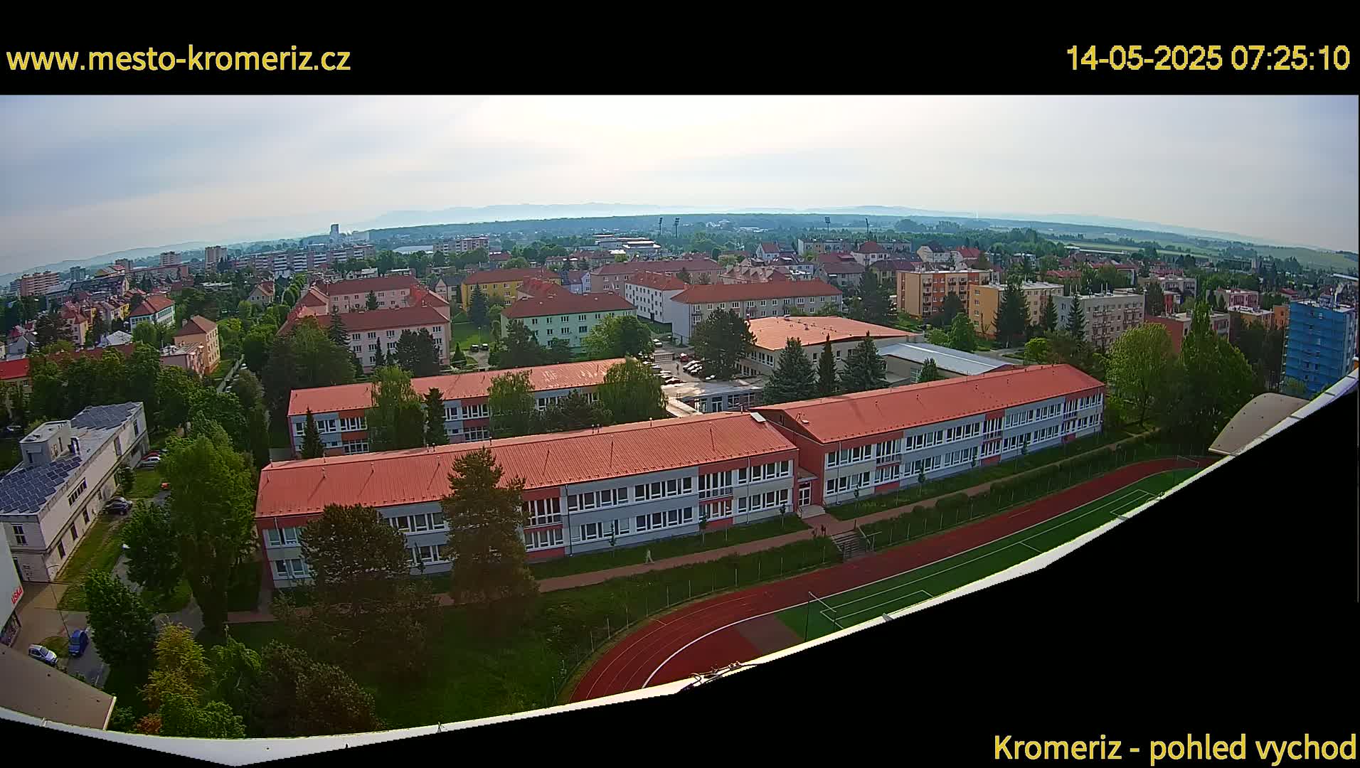 An aerial view shows a residential area with several buildings and a school with a running track, under a mostly clear sky.