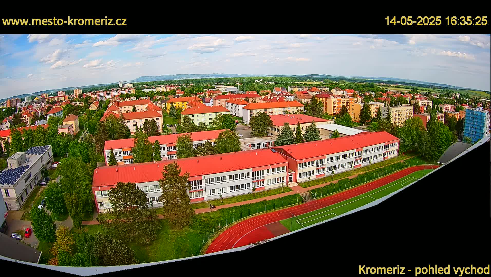 An aerial view shows a partly cloudy day over a residential area with a large school building and a running track, all surrounded by green trees.