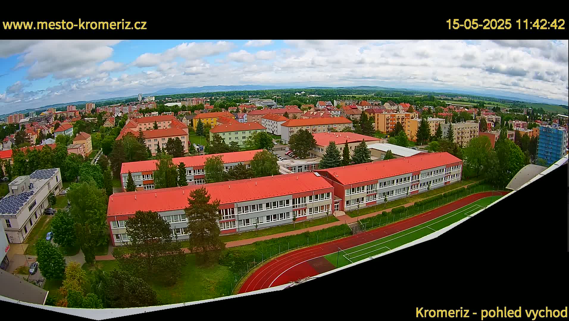 An aerial view shows a partly cloudy day over a town with several buildings, including a large school with red roofs and a running track, surrounded by green landscaping.