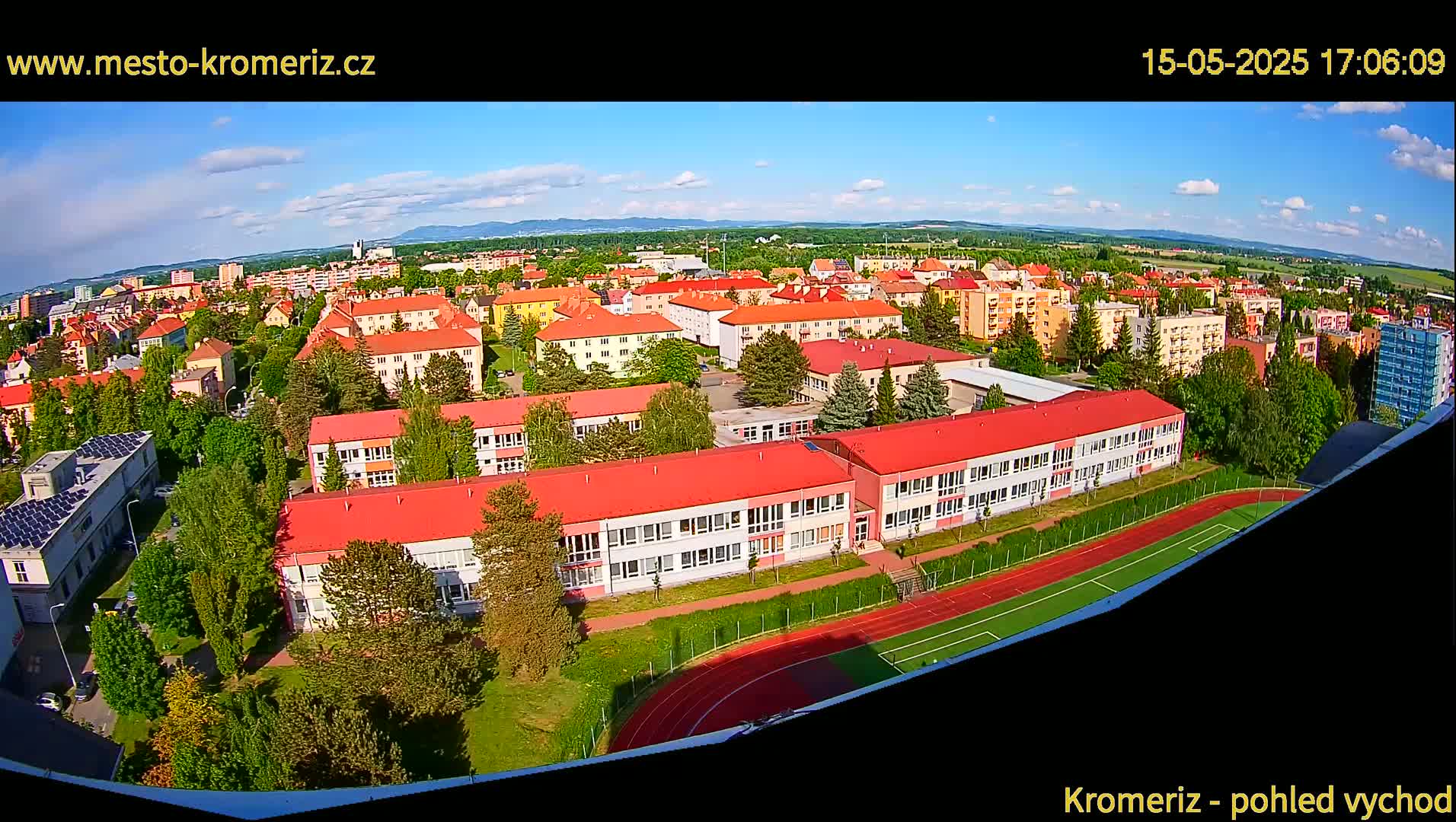 An aerial view shows a town under a partly cloudy sky, with a large school building and its surrounding athletic field prominently featured in the foreground.