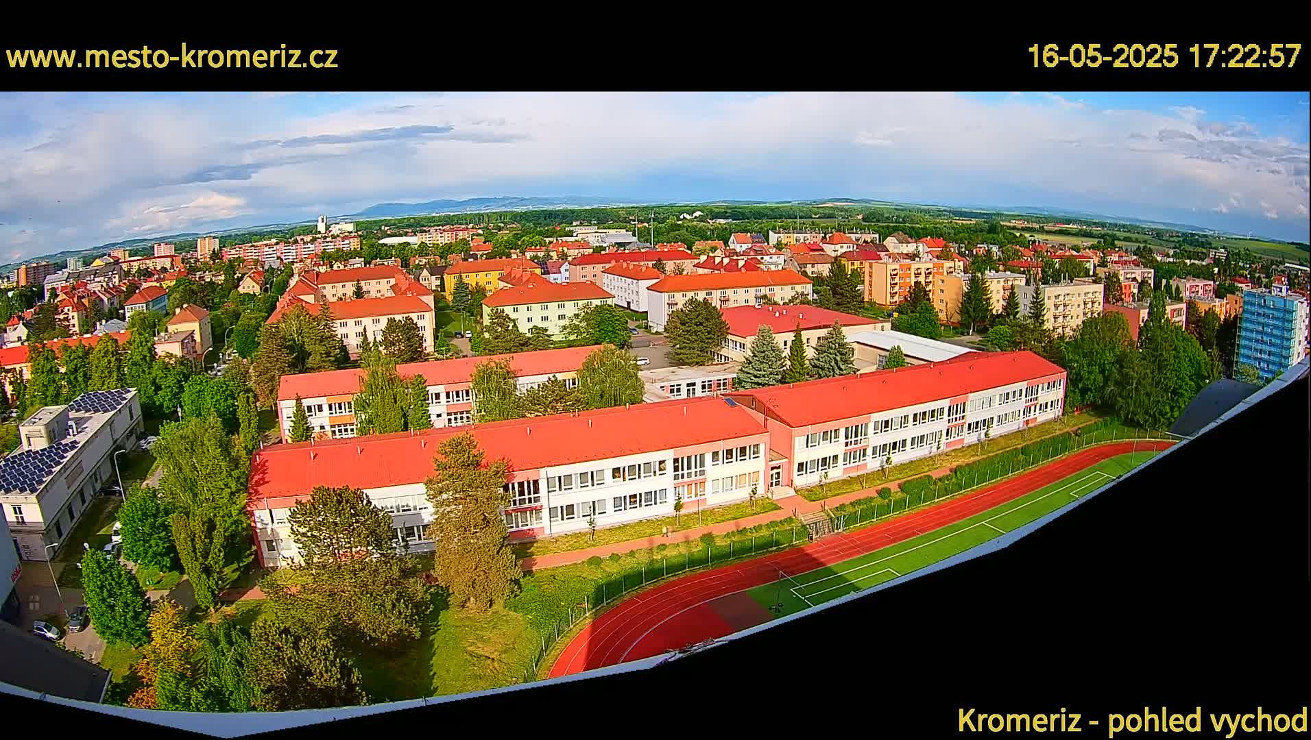 An aerial view of a town on a partly cloudy day shows a large school with red roofs and a running track, surrounded by other buildings and green spaces.