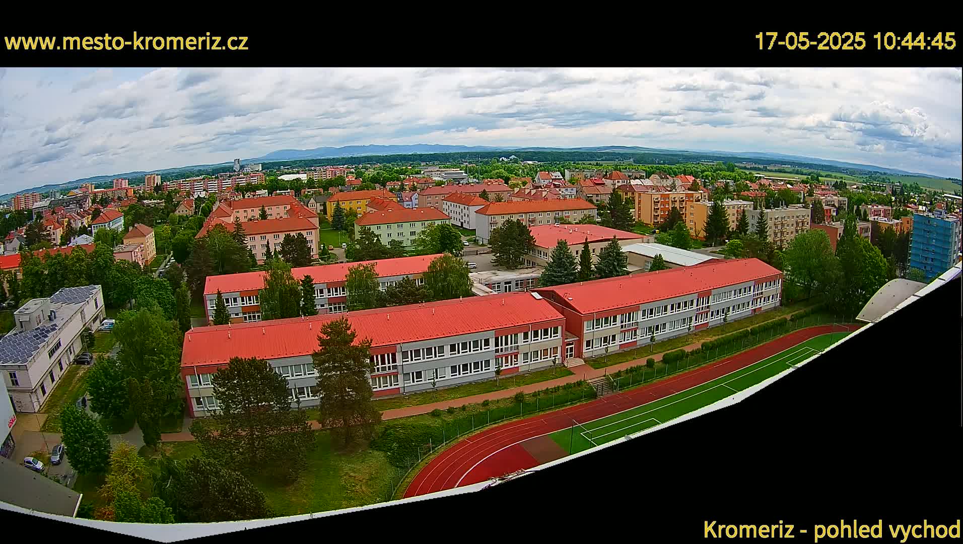 An aerial view shows a partly cloudy day over a town with numerous buildings, including a large school with a red roof and a running track, surrounded by green spaces.