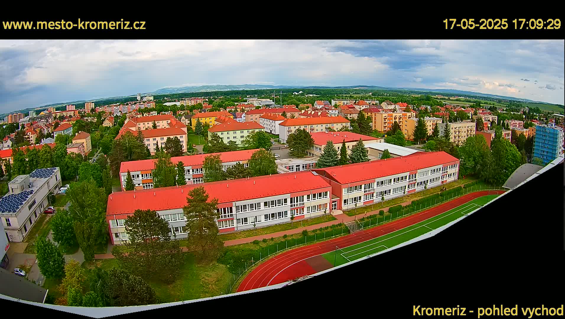 An aerial view shows a large school complex with red roofs and a running track, surrounded by residential buildings and green trees under partly cloudy skies.
