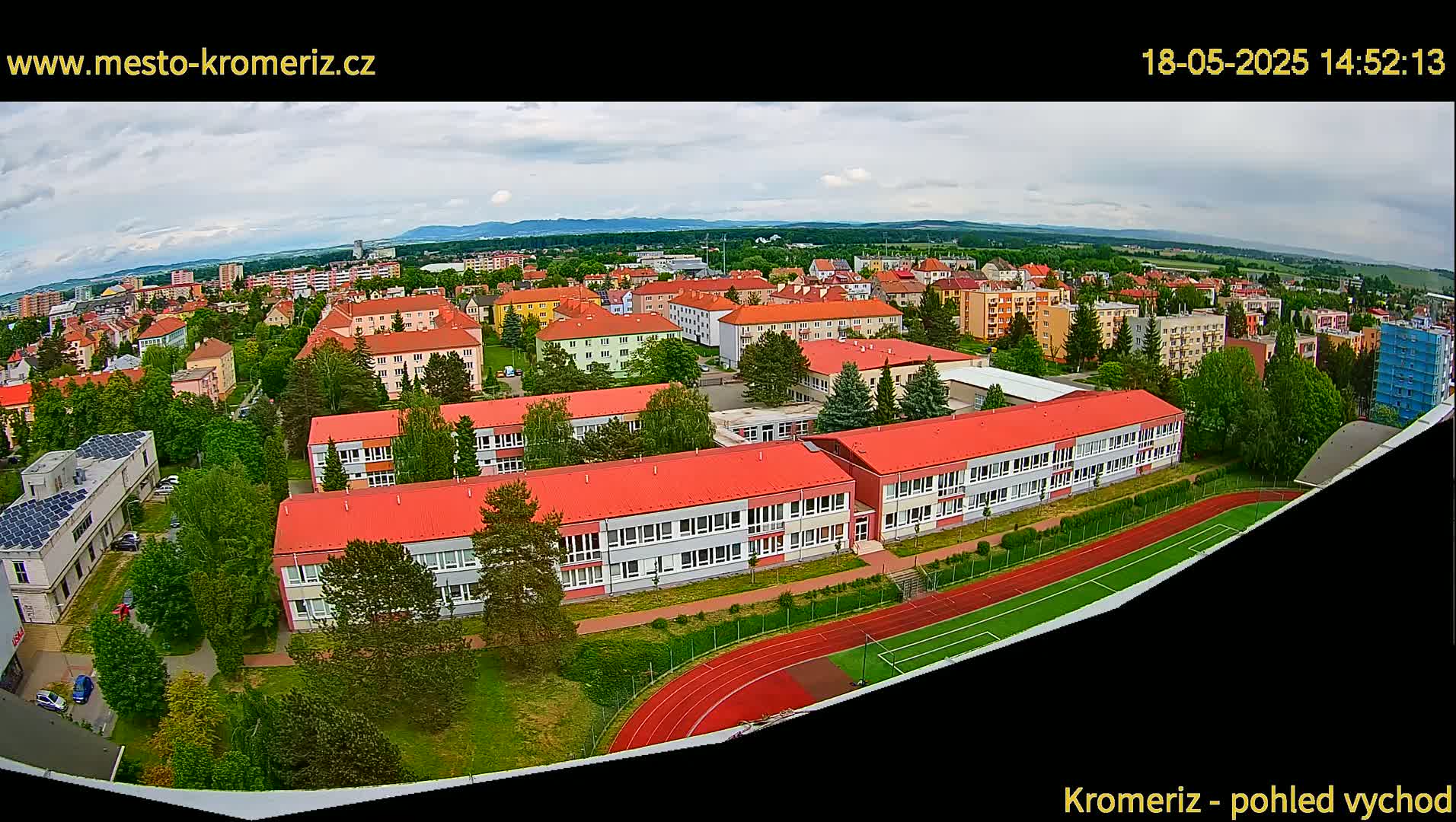 An aerial view shows a school with red roofs and a running track, surrounded by a residential area under partly cloudy skies.
