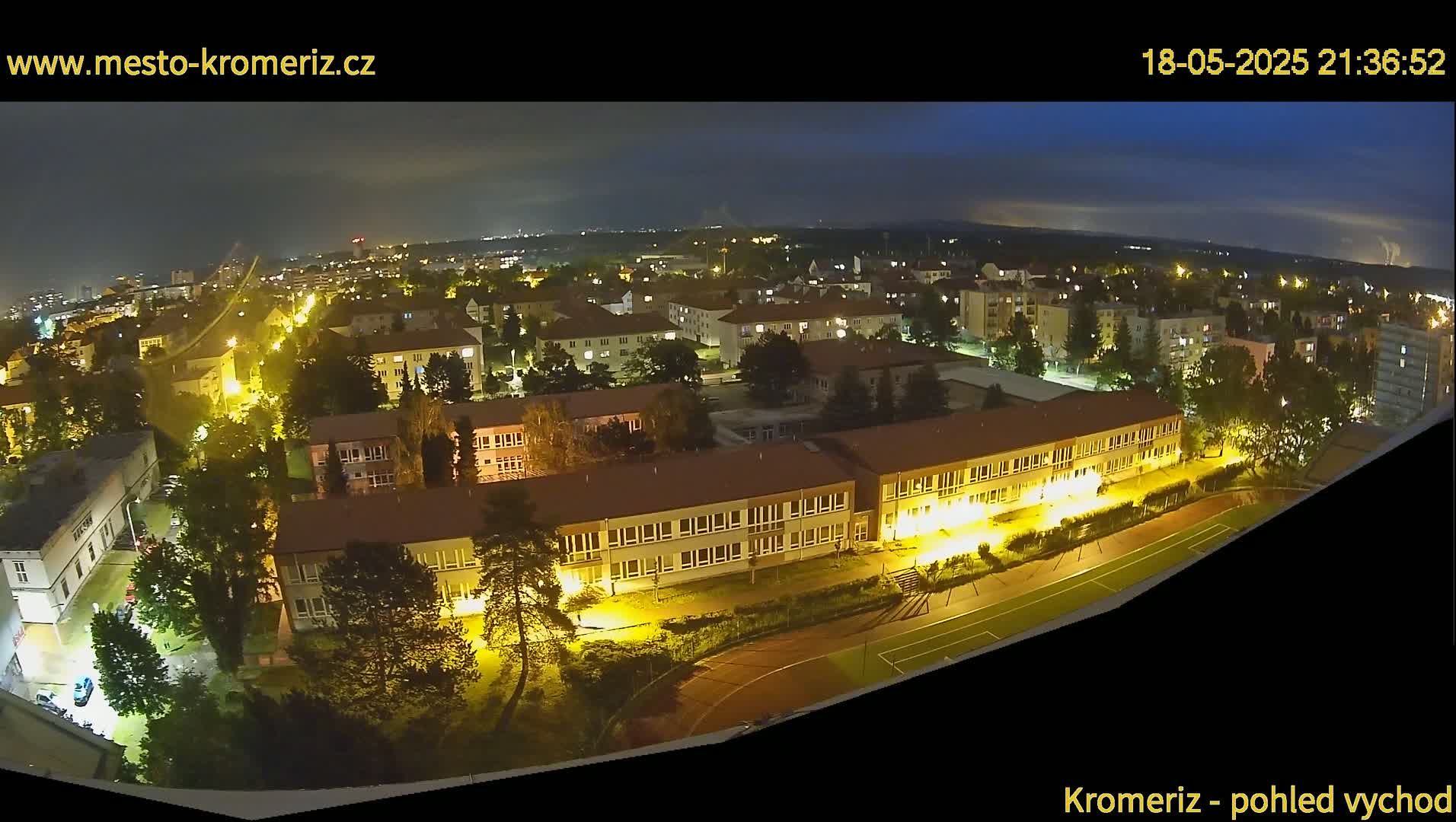 A nighttime, aerial view of a town, showing illuminated buildings and streets, with a large school building prominently featured, and a running track visible in the lower right.