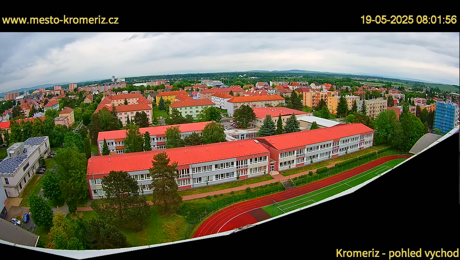 An aerial view shows a school with red roofs and a running track, surrounded by residential buildings under an overcast sky.